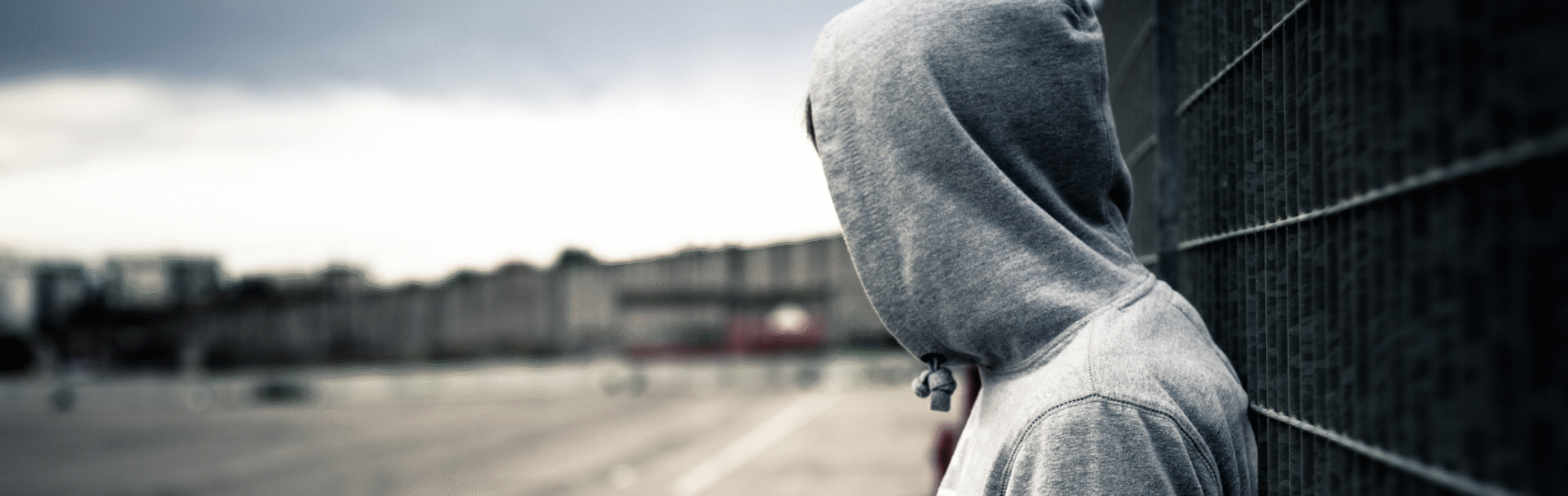 A youth wearing a light grey sweatshirt with the hood up is turned away from the camera and looking across an empty car park.
